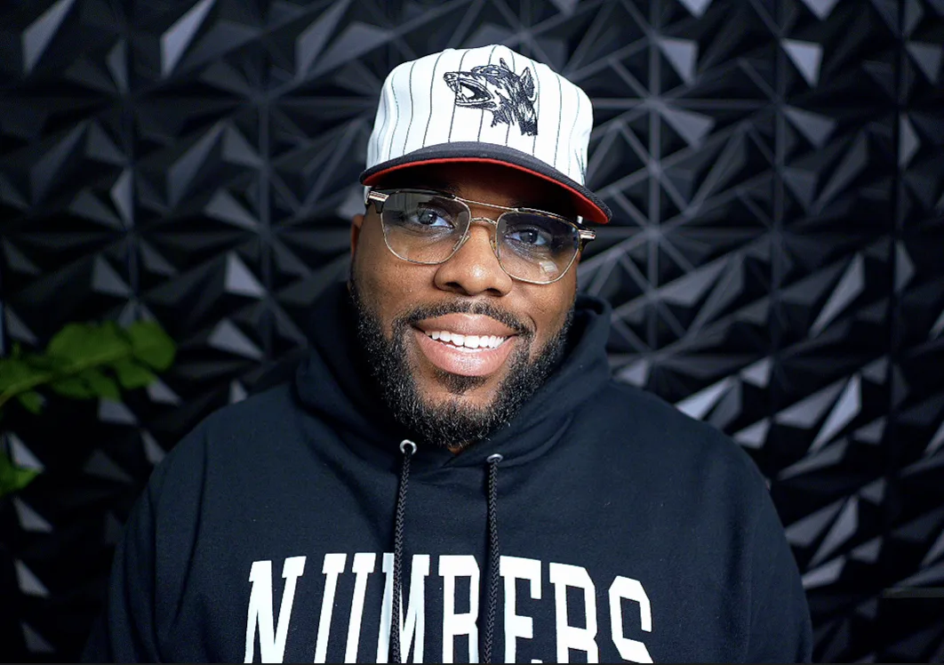Portrait of DeArcy “Trey” Scott smiling in a white cap and black hoodie against a dark geometric backdrop
